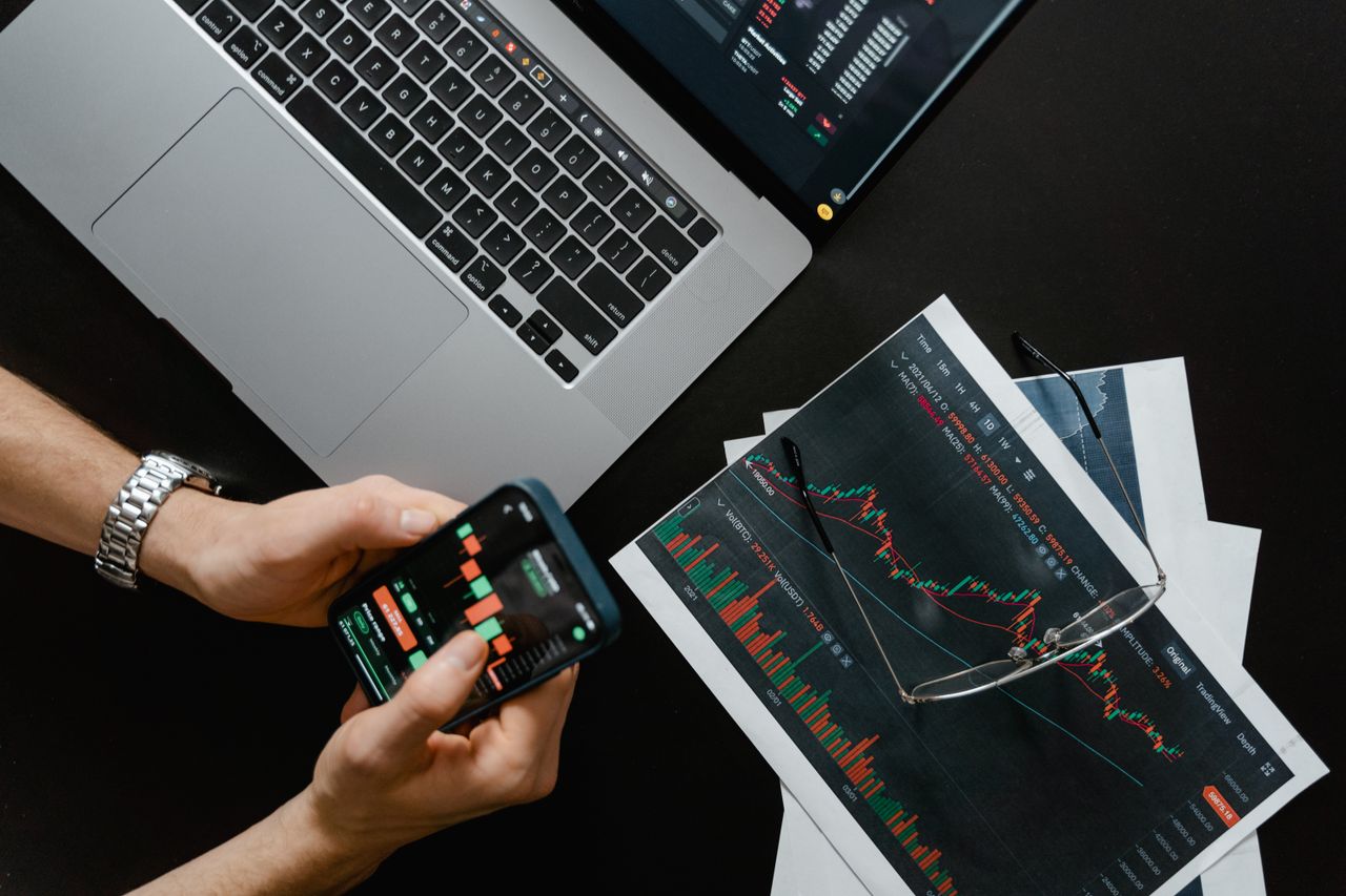 Des mains tenant un smartphone à côté d’un ordinateur portable affichant des graphiques boursiers, illustrant le trading numérique.